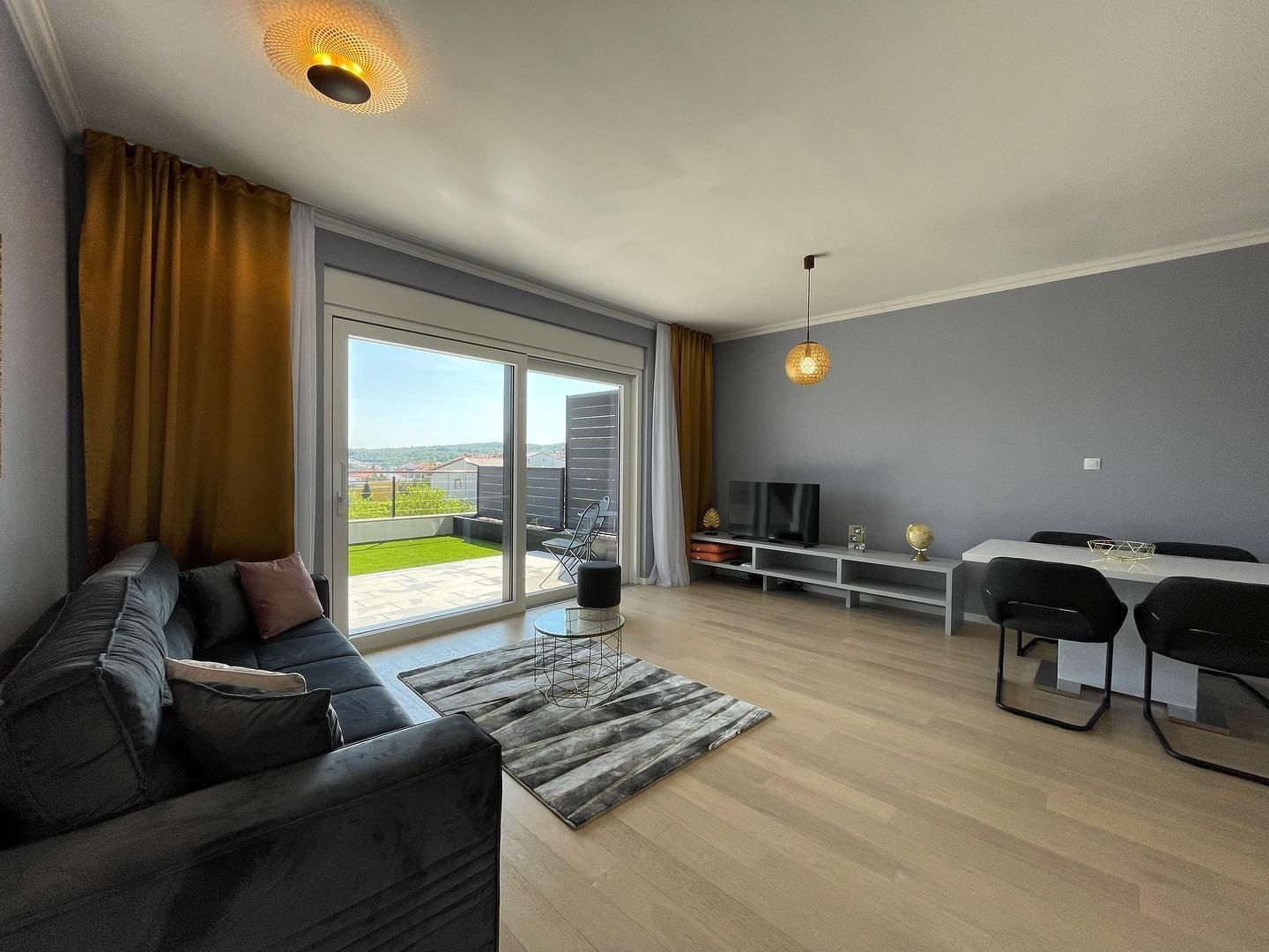 Wohnzimmer des Nanea Apartments: ein graues Samtsofa, ein weißer Esstisch und Glastüren, offen zu einer Rasenterrasse
